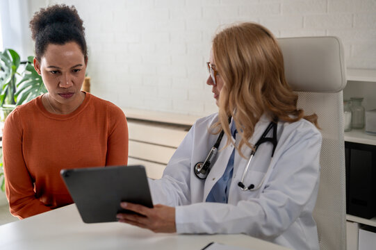Female doctor reviewing patient medical records on a clipboard while consulting a woman in a modern medical office. Physician studying health information during a routine appointment