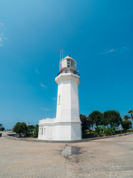 View of the historic Batumi Lighthouse standing against a clear blue sky, surrounded by lush trees and paved grounds in Batumi, Adjara, Georgia.