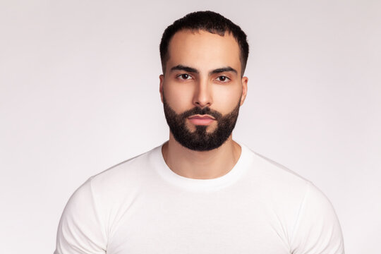 Serious bearded man wearing white t-shirt showing off groomed facial hair and smooth skin with calm expression professional male beauty grooming appearance isolated over gray background