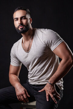 Confident guy in t-shirt sitting with hands on knees showing strong muscular arms wearing silver chain and bracelet in cinematic lighting isolated over black background