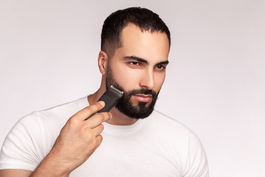 Bearded man using cordless clipper for beard styling maintaining sharp facial hair lines wearing white cotton t-shirt posing isolated over gray background