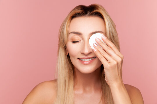 Blonde female removing eye makeup using soft cotton round during evening rejuvenation and hydration skincare procedure isolated on pink background