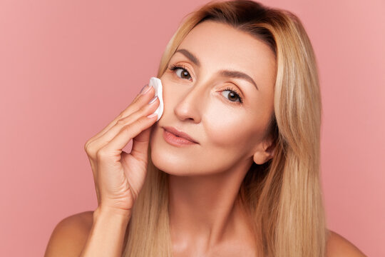 Beautiful lady using cosmetic cotton disk for face tonic application to achieve skin rejuvenation and healthy glow effect isolated on pink background