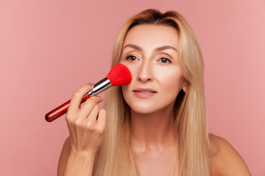 Mature blonde woman applying red powder brush on her face for professional makeup routine and skin perfection isolated on pink background