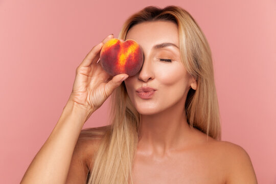 Joyful blonde woman holding peach over her eye and making kiss face, enjoying summer fruit benefits for daily skincare isolated on pink background