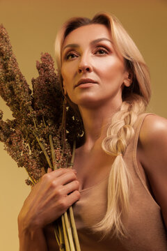 Graceful woman looking up and holding floral bunch of dried weeds while wearing beige top and posing for artistic fashion portrait isolated over yellow background