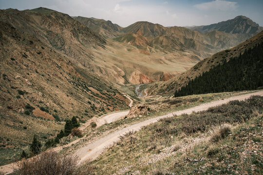 View of a winding dirt road through the rugged mountainous landscape of the Tian Shan range with sparse vegetation and rocky slopes in Naryn Region, Kyrgyzstan.