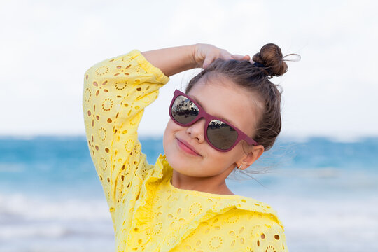 Smiling young girl in a yellow eyelet dress poses with sunglasses at the beach, enjoying a sunny summer day and relaxed, carefree seaside mood