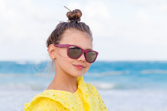 Smiling girl in a bright yellow dress wears purple sunglasses on a sunny beach, posing against a soft-focus ocean backdrop that conveys summer fun, relaxation and carefree vacation vibes