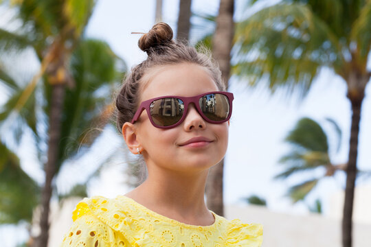 Confident little girl in a yellow eyelet dress smiles wearing sunglasses against swaying palm trees on a sunny tropical vacation, conveying summer, joy, leisure and carefree childhood moments
