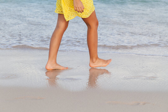 Barefoot girl walking along the shoreline in a yellow summer dress, leaving soft reflections on wet sand as gentle waves lap the shore, conveying relaxation, freedom and easy vacation vibes