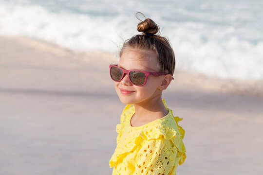 Smiling little girl wearing pink sunglasses and a yellow eyelet dress stands on a sunny sandy beach, looking toward the sea with gentle waves and a carefree summer vacation mood