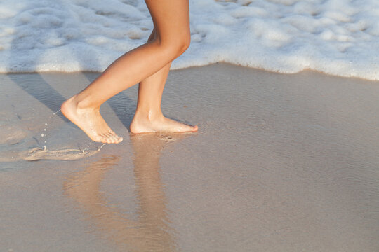 A girl walks barefoot along the wet sandy shore as gentle waves wash over her feet, casting a warm reflection and evoking relaxation, freedom and a peaceful summer coastal mood