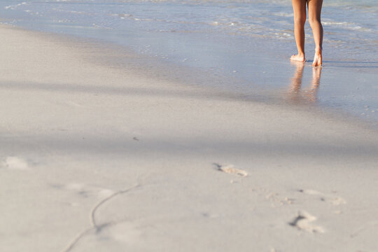 Barefoot woman walking along a sandy shoreline with wet sand reflections, soft waves and footprints. Quiet summer beach scene conveying relaxation, solitude, escape and peaceful coastal atmosphere
