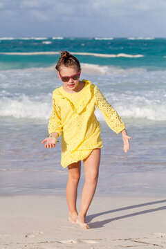 A young girl in a bright yellow dress and sunglasses walks along a sunlit sandy beach with turquoise waves behind her, evoking playful summer joy, carefree childhood moments and seaside vacation vibes