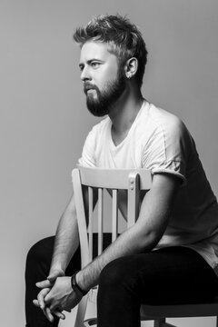 Monochrome shot of serious bearded man sitting on chair with clasped hands looking away in profile isolated on gray background
