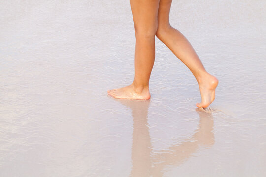 Barefoot person walking along wet sand at the shoreline, feet touching shallow water and creating soft reflections; evokes calm, relaxation and the freedom of a peaceful summer seaside stroll