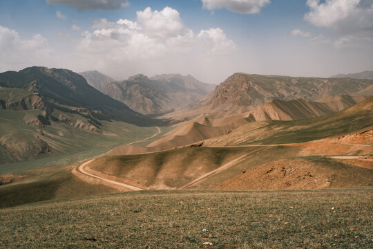 View of Song-Kul landscape with rolling hills, a winding dirt road, and distant mountains under a cloudy sky in Naryn Region, Kyrgyzstan.