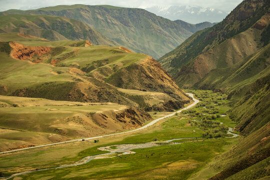 View of the vast landscape of Song-Kul with rolling green hills, a winding road, and a meandering river flowing through the valley in Naryn Region, Kyrgyzstan.