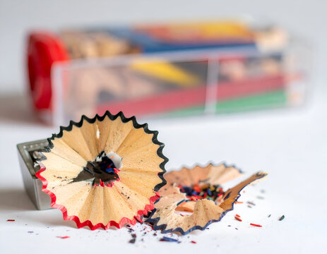 A metal pencil sharpener with fresh wood shavings and graphite dust on a white surface, with a blurred plastic container of colorful pencils in the back.