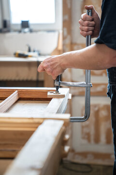 Craftsman at workbench in restoration workshop securing parts of old wooden window frame using clamps, skilled carpenter carefully fixating wood detail with clamp instrument, prepares for restoration.