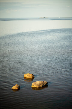 Three rocks in calm shallow sea water illuminated by warm sunset light, smooth surface with almost no waves, distant island on horizon, peaceful seascape, quiet evening atmosphere and still water.