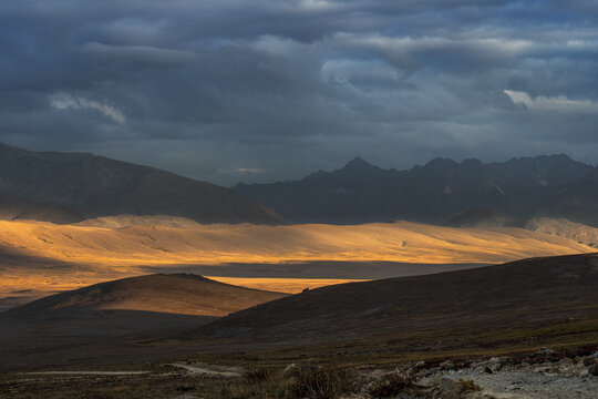 View of Deosai National Park with golden sunlight illuminating the rolling hills under a dramatic dark cloudy sky in Skardu, Pakistan.