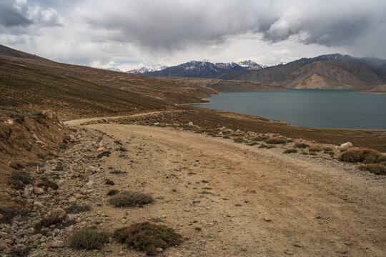 View of a dirt road winding through the arid landscape towards a lake in the Pamir Mountains, Jirgatol, Gorno-Badakhshan Autonomous Region, Tajikistan.