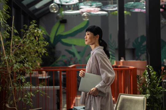 Asian businesswoman holding laptop looking away in modern office