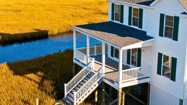A raised coastal cottage in Georgia's Golden Isles, Tybee Island, with white clapboard siding, dark green shutters, harsh sun casting sharp shadows across a wraparound porch and marsh grass.