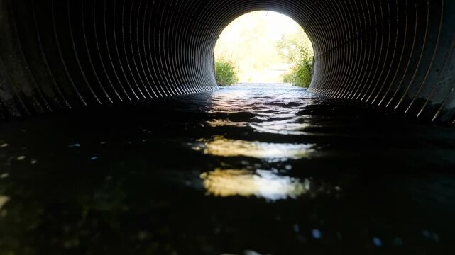 Pushing by current, stream water forming ripples toward camera in corrugated culvert, with sunlight