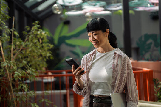 Asian woman smiling using smartphone holding laptop
