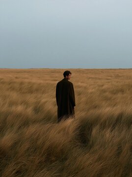Man standing in the middle of a vast field of tall, golden grass. he is wearing a long black robe and appears to be deep in thought.