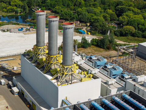 Aerial view of a power plant featuring three tall industrial chimneys with yellow support structures, surrounded by industrial infrastructure and lush green trees in the United Kingdom.