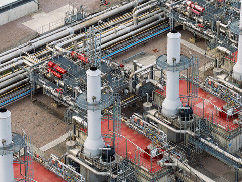 Aerial view of an industrial facility featuring complex networks of metallic pipes, tall white chimneys, and red-painted service platforms, United Kingdom.