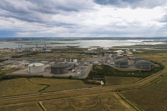 Aerial view of an industrial facility featuring large cylindrical storage tanks situated near a coastal estuary with surrounding grassy fields and a cloudy sky in the United Kingdom.