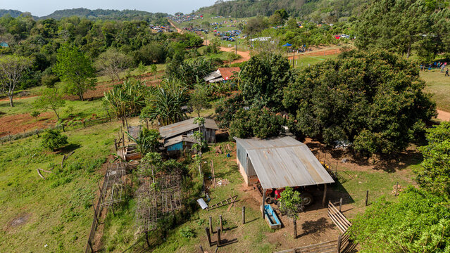 Aerial view of a rural farmstead with metal-roofed buildings and lush greenery, with a distant gathering of vehicles and tents, Nueva Alborada, Itapua Department, Paraguay.