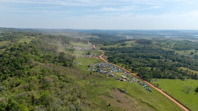 Aerial view of the Rally Paraguay event with spectators and tents gathered along a winding dirt road in the green hills of Nueva Alborada, Itapua Department, Paraguay.