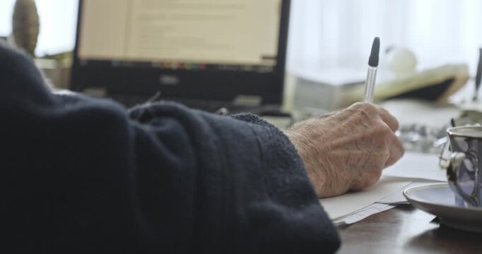 Senior person writes a traditional letter at home beside a laptop, contrasting manual correspondence with modern digital communication.