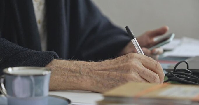 Close-up of an elderly person's hands simultaneously taking handwritten notes and operating a mobile smartphone at a desk.