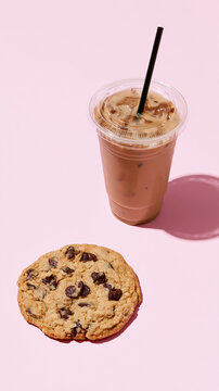 Iced coffee with milk in a takeaway cup beside a chocolate chip cookie on pastel pink background, refreshing caf? drink and sweet snack with copy space