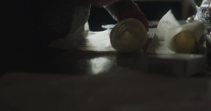 Person in a low-lit room rolls large paper sheets across a table, creating an atmospheric scene suggestive of archive research, documents or architectural plans.