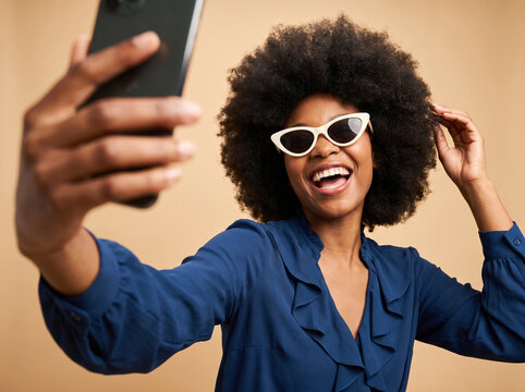 Happy young Black woman with an afro taking a selfie with her smartphone, wearing stylish white sunglasses and a blue shirt.