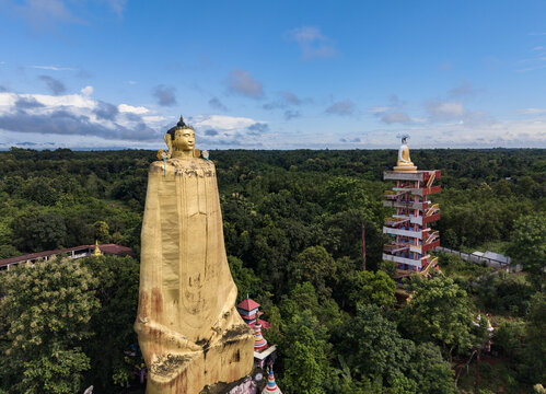 Aerial view of a large golden Buddha statue and a multi-tiered pagoda structure surrounded by lush green forest under a blue sky in Khagrachari District, Chittagong Division, Bangladesh.