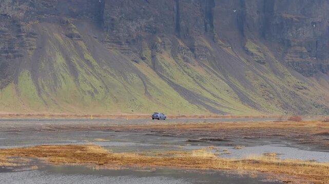 アイスランドの断崖の下を走る一台の車、湿原と苔むした山肌（Car Driving Along Cliff Base in Iceland）