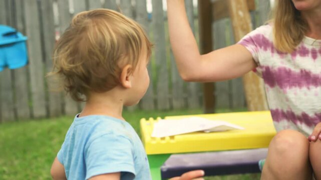 Toddler approaching mother folding paper airplanes by backyard fence