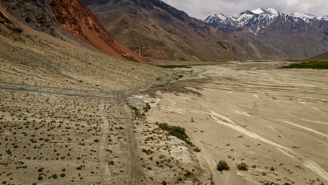 Aerial view of the Bartang River valley featuring a dry, rocky riverbed, rugged mountain slopes, and snow-capped peaks under an overcast sky in Chah Ab, Gorno-Badakhshan, Tajikistan.