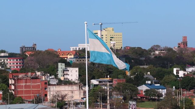 Argentine national flag waves above the Posadas skyline from the historic El Mastil flagpole, with urban buildings, trees and construction cranes visible across the city, slow motion.