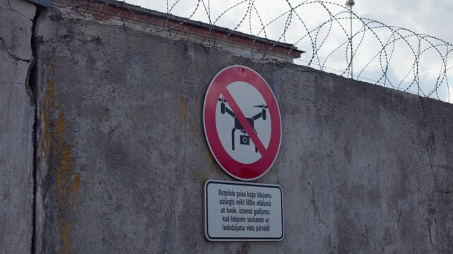 Close view of official Latvian language warning sign prohibiting drone flights on concrete perimeter fence with barbed wire at Daugavpils correctional site.