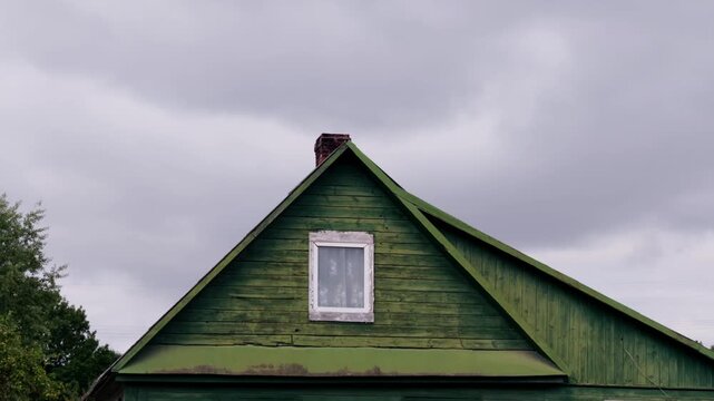 Low angle static shot shows triangular wooden gable roof and window of old green painted house against cloudy sky in Daugavpils, Latgale region, Latvia.